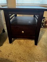 Front view of one dark brown wooden end table showing drawer with metal knob and open shelf.