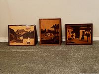 Three wood marquetry pictures displayed on a floor against a wall, showing front view of scenic images in various brown wood tones.