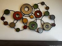 Front view of metal wall art showing circular shapes in various colors connected by black metal rods, mounted on a cream wall above a dark wood dresser.