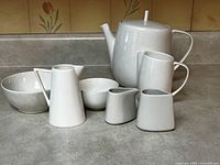 Photo showing full set of white porcelain and bone china items including teapot, creamer, and bowls placed on counter.