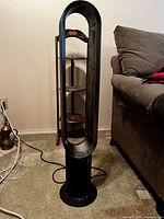 Front view of Dyson bladeless tower fan placed on carpeted floor near couch and an adjacent shelf unit.