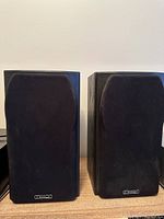 Front view of two black Mission loudspeakers with black fabric grilles and Mission logos at the bottom of each grille, placed side by side on a wooden surface.