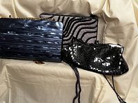 Three women's evening clutches arranged side by side on cream fabric background. Shiny blue clutch, grey fabric clutch with bead swirls, and black sequin clutch shown.