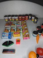 Photo showing multiple miniature canned goods including apple sauce, tomato sauce, and others along with boxed food items and small plastic foods.