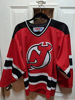 Front view of red New Jersey Devils hockey jersey on hanger, showing team logo and striping details.