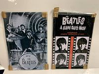 Photo of two new Beatles tin signs in original packaging taped at corners, one with black and white band photo with instruments and Ludwig drum logo, the other with stylized portraits and A Hard Day's Night soundtrack text