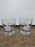 Two clear glasses on a brown surface showing the Toronto Maple Leafs logo etched on each.