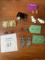 Overview photo showing all animal-themed earrings including panther face, fish, cat, dog, owl, butterfly, and deer earrings on a wooden surface.