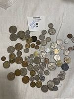 Top-down photo showing an assortment of approximately 70 international coins of various sizes and colors laid out on a white fabric surface.