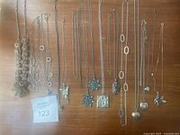 Top down image showing 14 silver-tone necklaces laid out on a wooden surface. Different chain designs and pendants including heart-shaped lockets and floral pendants visible.