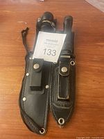 Two vintage camping knives side by side in their black leather sheaths on wooden surface