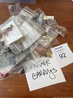 Photo of multiple small plastic bags containing numerous pairs of vintage earrings, some on cards, on wooden surface along with a handwritten label 'VINTAGE EARRINGS' and a MaxSold tag 92.