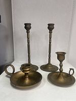 Four brass candlesticks: two tall and slender with rounded bases, two shorter chamberstick style with circular handles, all with a patina consistent with light use.