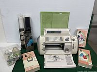 Front facing photo showing Cricut machine, font cartridges, green paper mats, vinyl packages, and paper punch tools arranged on green table.