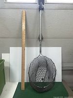 Photo of large fishing net stood upright next to a wooden ruler for size reference. Net has aluminum handle and black rubber mesh netting.