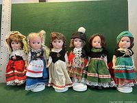 Photo showing six foreign country dolls lined up against green backdrop with ruler for scale.
