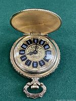 Gold tone pocket watch open showing black Roman numeral hour markers and intricate floral pattern on dial and case.