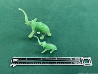 Two miniature jade elephant figurines placed on green surface beside a ruler for size reference.