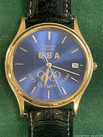 Front view of Seiko Olympic member watch with blue dial, gold-tone case, and black leather strap showing USA and 1992 Olympic Team logo.