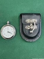 Front view of silver-tone pocket watch next to black leather case on green background.
