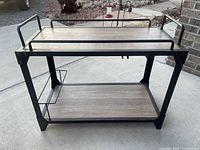Overall front and side view of the rustic bar cart with two wood shelves and black metal frame, showing handles and the metal rack on the side shelf.