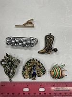 Top view of six vintage brooches arranged on a white surface with a pink ruler for scale, showing front side details including enamel, rhinestones, and pearl embellishments.