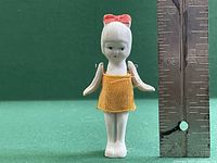Front view of the vintage miniature porcelain doll, showing its painted face, arms extended slightly outwards, orange fabric dress, and pink bow on the head. A ruler placed beside the doll shows it is about 2.5 inches tall.