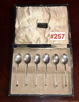 Open presentation box lined with satin containing seven coffee spoons arranged in fitted velvet slots.