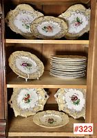Three plates displayed on a wooden cabinet shelf showing scalloped edges and gilded border.