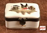 Front view of white porcelain box with painted hummingbird and foliage on lid, metal hinge at front