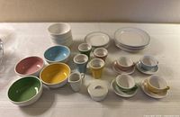 Overview of miniature ceramic tea set components including cups, saucers, bowls, plates, creamer, and sugar bowl on a white cloth surface.