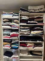 Full view of two white shelving units packed with neatly folded knitwear, shirts, and pants.