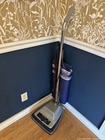 Sanitaire heavy duty upright vacuum cleaner with silver base, black accents and blue fabric bag standing against a navy blue and beige wallpapered wall with wooden floor.