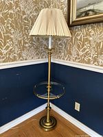 Full view of vintage brass floor lamp with pleated cream lampshade and round glass tray table in corner.