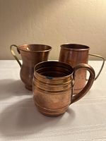 Three vintage copper mugs with different shapes and brass handles shown together on a white cloth surface.