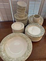 Overall view of stacked plates, bowls, and cups showing cream ceramic with raised green vineyard and grape designs around edges.