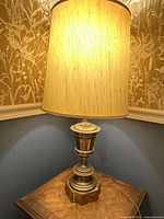 Full view of the brass table lamp turned on, showing the beige fabric shade and the lamp base on a wooden table against a blue wall with floral wallpaper above.