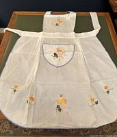 Full apron laid flat showing white fabric with floral embroidery at hem, bib, and pocket, and long tie straps