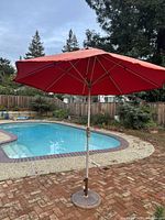 Full view of the red patio umbrella set up outdoors near a pool, mounted in a round metal weighted base on a brick patio.
