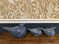 Front view of set of three vintage blue stoneware bird figurines in varying sizes on wooden surface against floral wallpaper.