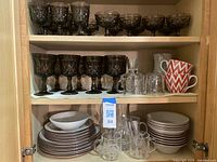 Cabinet shelf displaying amber faceted wine glasses along with clear glass mugs, decanter, and red and white ceramic mugs.
