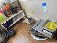Wide view of kitchenware items stacked on floor and shelf including pots, pans, baking trays, citrus juicer, and plastic items.