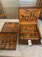 Full view of two open wooden cases with multiple bronze flatware pieces arranged in rows secured in slots, with some utensils wrapped in plastic.