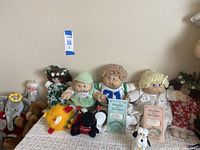 Wide shot showing three Cabbage Patch Kids dolls in center among various other stuffed animals, including elephant, lion, and small dog plushes.