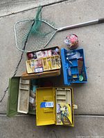 Overview of fishing gear set laid out on concrete including three tackle boxes with fishing gear, a fishing net, and extra accessories.