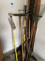 Full view of assorted garden and yard tools standing against wall, showing various rakes, shovels, and cultivators with wood and metal handles.
