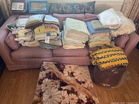 Photo showing a large assortment of folded towels, blankets, linens, and cushions arranged on a pink couch and on the floor in front. The textiles vary in color and pattern, mostly cotton fabric.