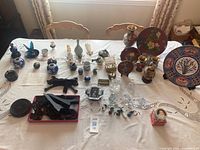 Table display showing the entire collection of ceramic and metal decorative items including plates, vases, bowls, figurines, and small trinkets, arranged on a white embroidered tablecloth.