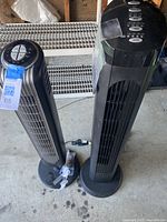 Two black tower fans standing side by side on a concrete floor, one taller and wider, both show dust and wear