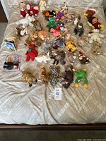 Wide top-down view showing all plush animals laid out on a bed, displaying the wide variety of colorful animals and the lot number card.